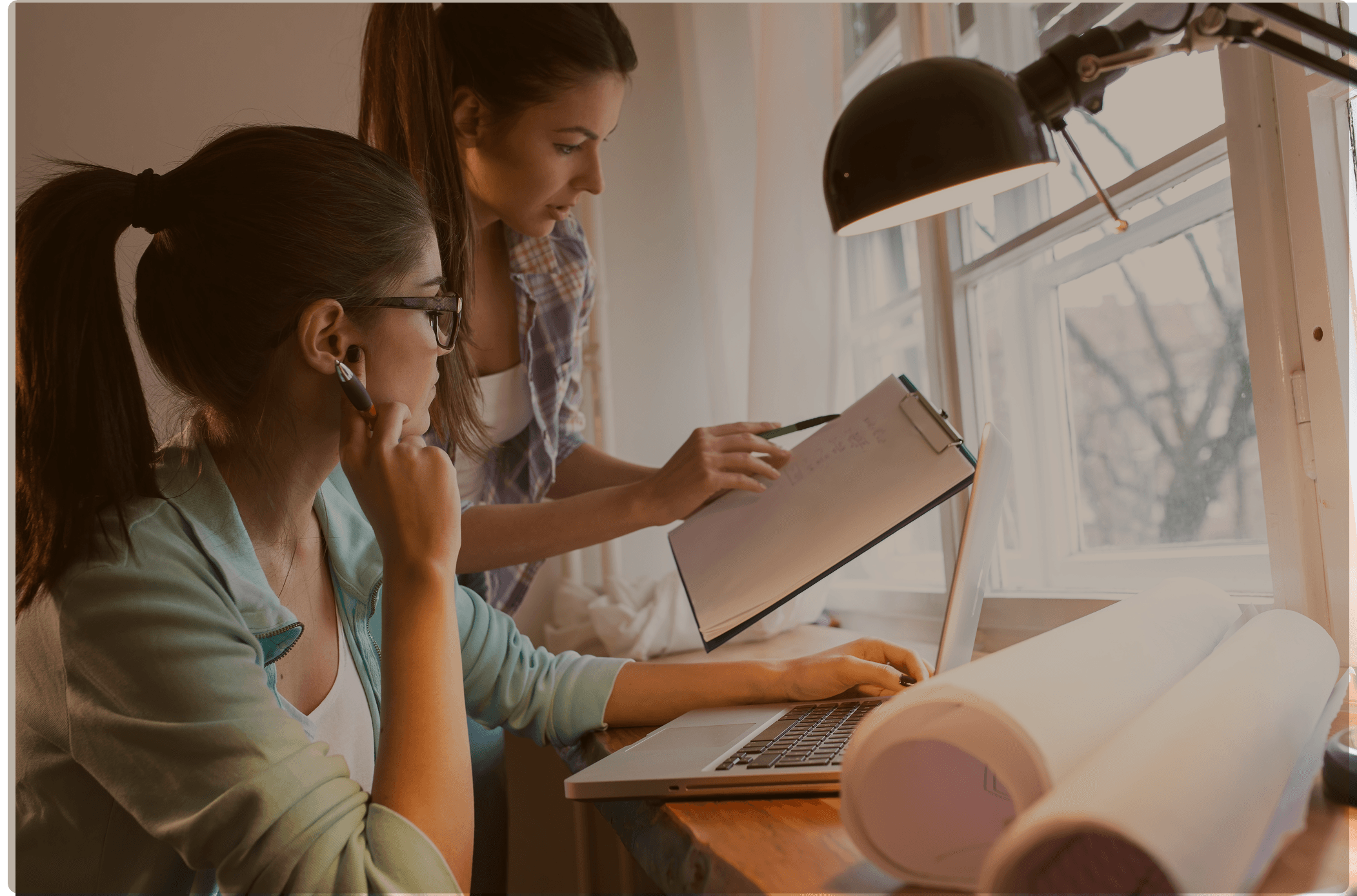 Two women collaborating at a desk by a window, one taking notes on a clipboard while the other works on a laptop, with rolled blueprints nearby.