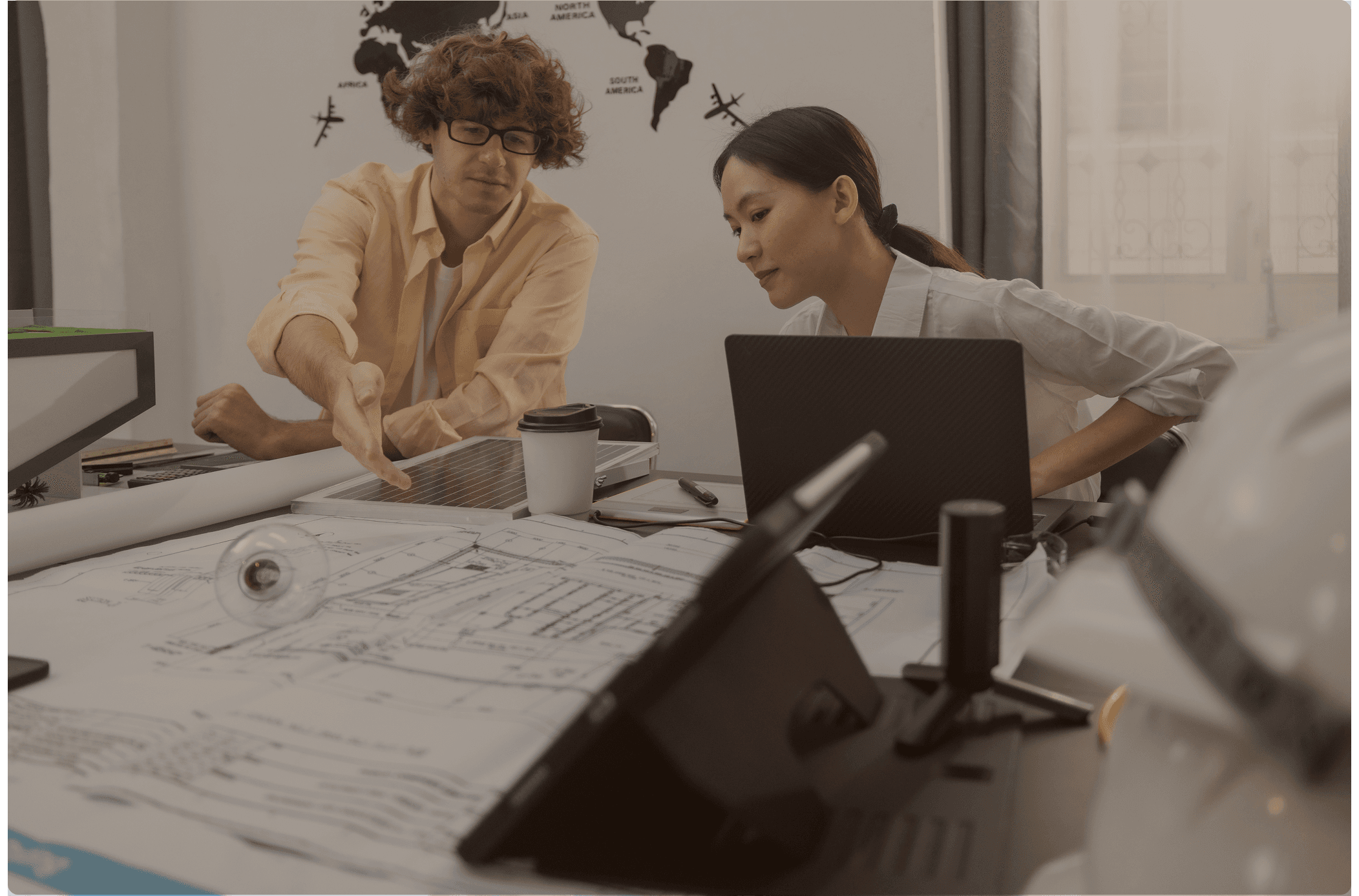 Man and woman reviewing architectural blueprints and a solar panel at a desk, with a laptop, coffee cup, and world map on the wall behind them.